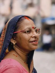 An elegant woman with glasses and traditional clothing gracefully smiles against a blurred background.